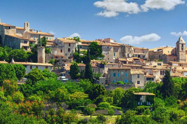 A charming hilltop village perched on elevated terrain, featuring traditional stone buildings with terracotta roofs and a prominent church tower. The settlement is nestled among lush green vegetation under a brilliant blue sky dotted with white clouds, creating a quintessential Mediterranean countryside scene that exemplifies timeless rural architecture and natural beauty.